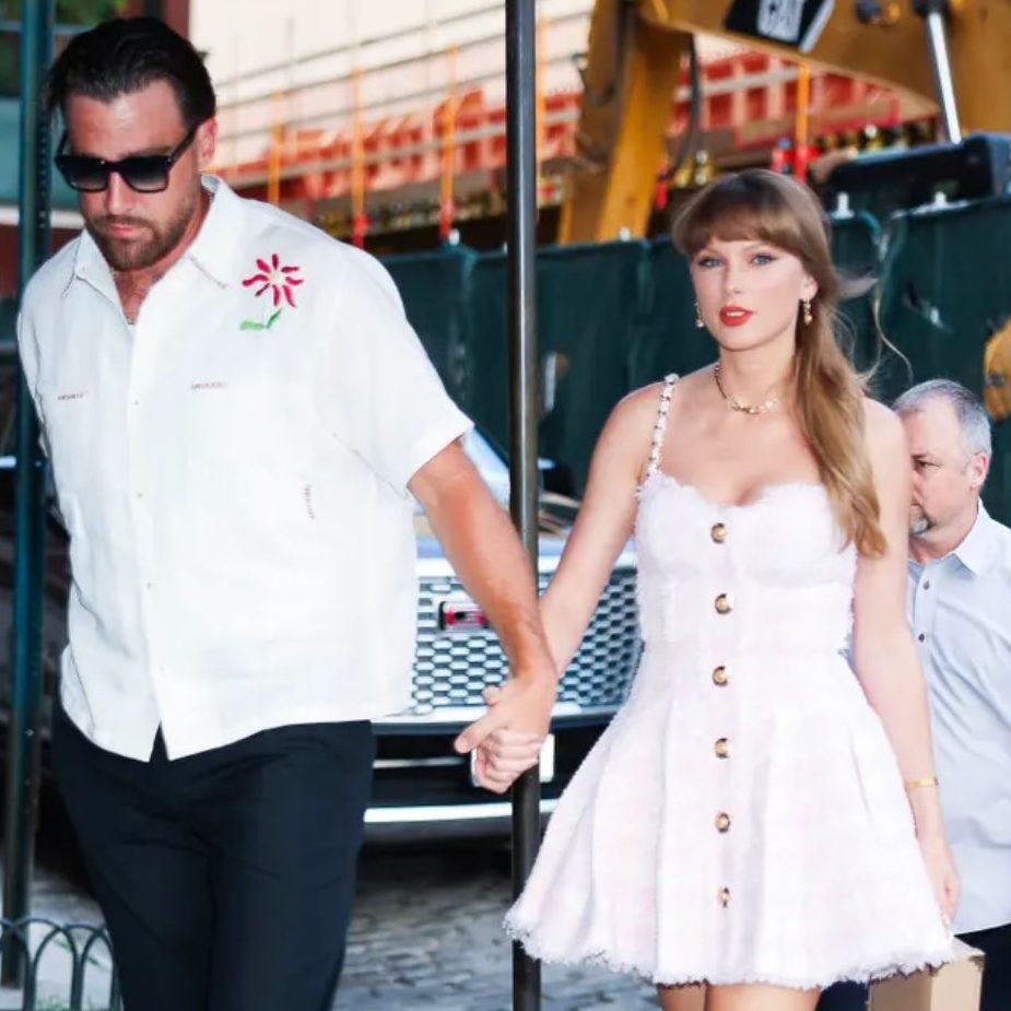 Taylor Swift wearing a pink and white houndstooth dress with beige colored heels and Travis Kelce in a white shirt and black pants holding hands walking into a restaurant in New York City